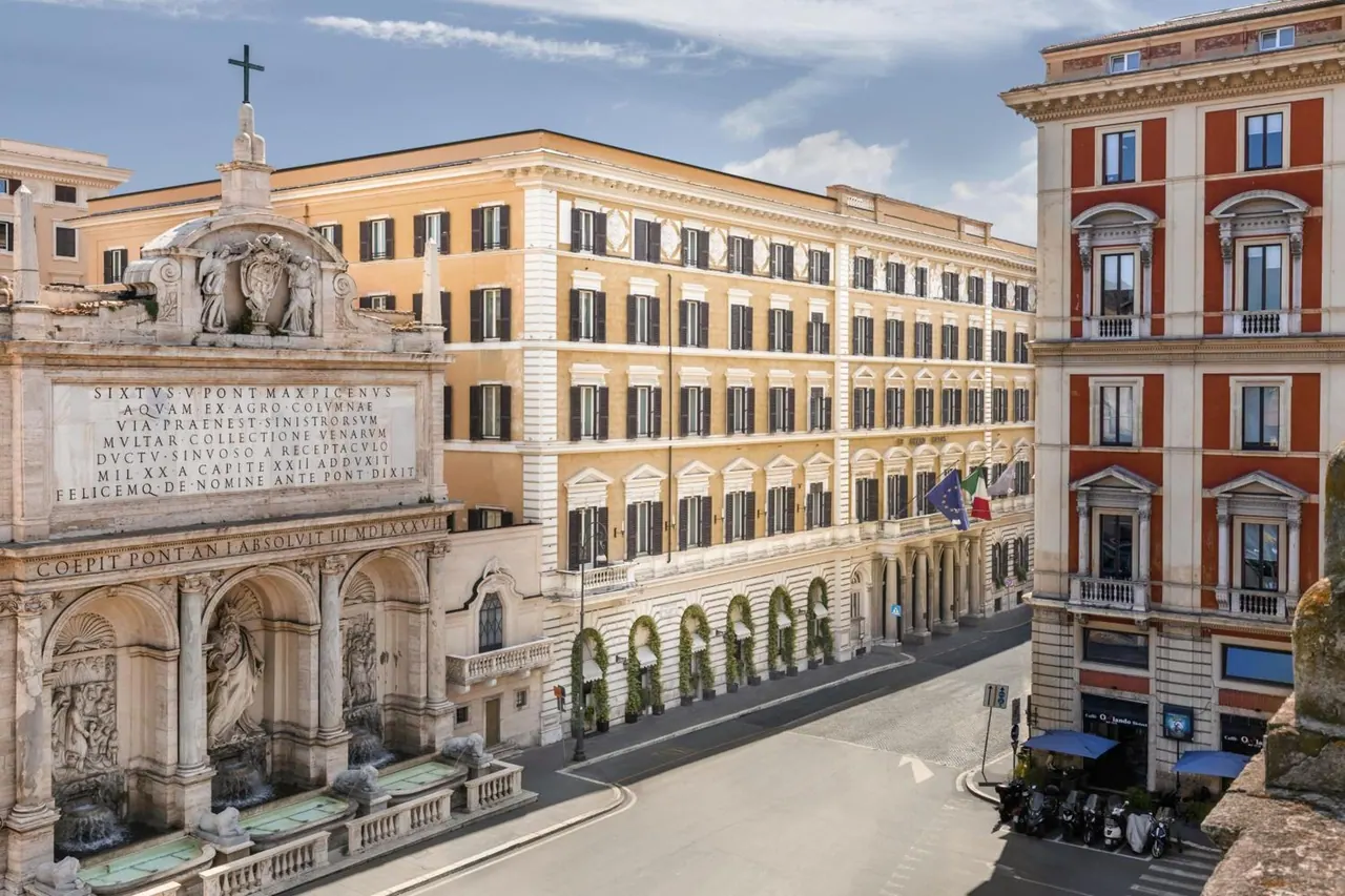 Exterior view of a cream and orange hotel building on a Rome street, with the Fontana dell'Acqua Felice fountain at left and parked scooters at lower right.