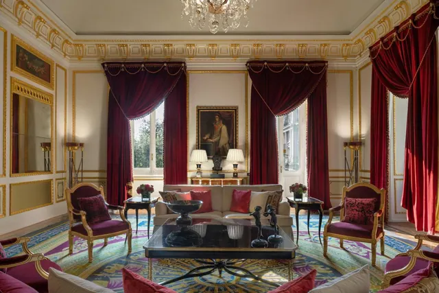 A formal living room with red velvet curtains, gold-framed armchairs, a sofa, a glass coffee table and a crystal chandelier.