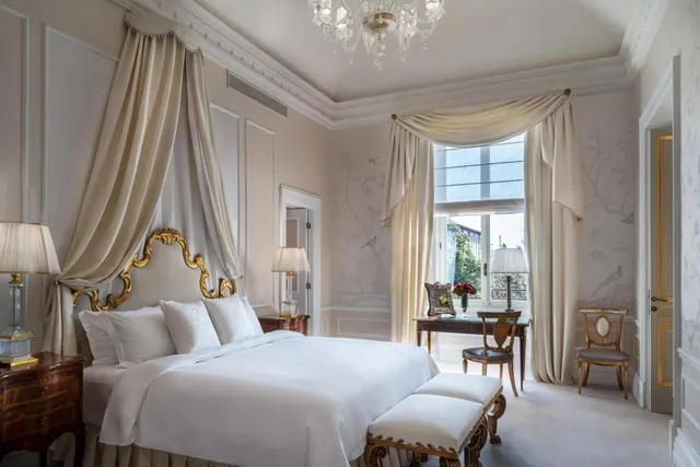 A bedroom with a canopied king bed with a gilt headboard, a chandelier overhead, and a writing desk by a tall window with cream drapes.