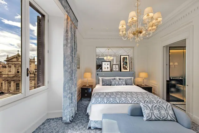 A bedroom with a double bed, a glass chandelier, and a tall window looking out onto a historic church facade.