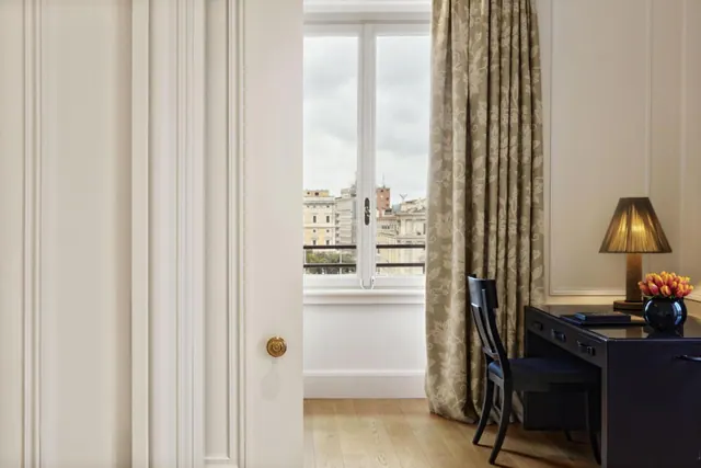 A cream-panelled hotel room with a partly open door, a window onto city buildings under a cloudy sky, and a dark desk with a lamp and tulips.