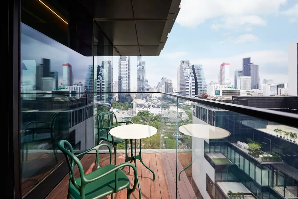 A wooden balcony with green chairs and two round white tables overlooks a city skyline of high-rise towers under a partly cloudy sky.