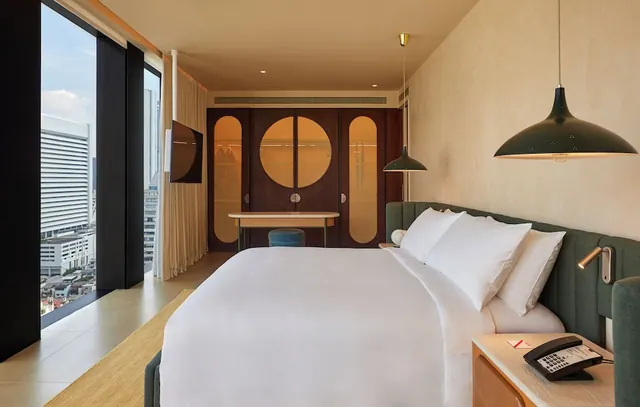 A bedroom with a made king bed, a wall-mounted television, a wooden wardrobe with circular cutouts, and floor-to-ceiling windows looking onto city buildings.