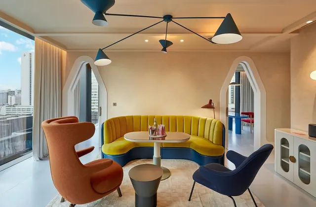 A living area with a curved yellow sofa, two armchairs, a round table and a city view through large windows on the left.