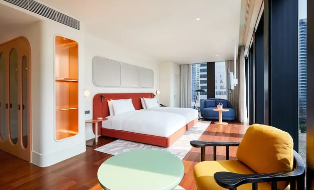 A bedroom with a large bed with a red-orange headboard, a blue armchair and a yellow chair beside floor-to-ceiling windows that look out over high-rise buildings.