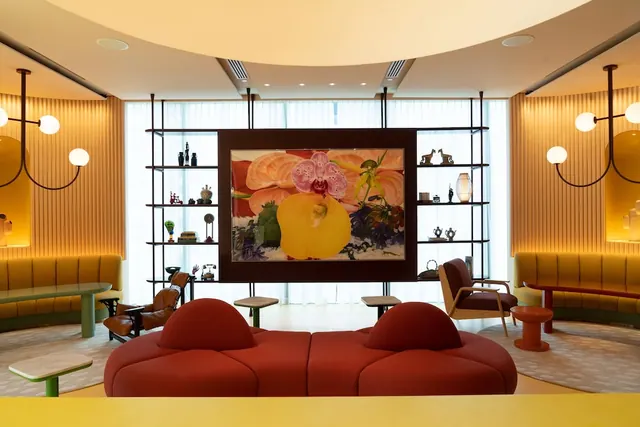 A lounge seating area with a red curved sofa, yellow banquettes and a large framed flower painting in front of tall windows with sheer curtains.