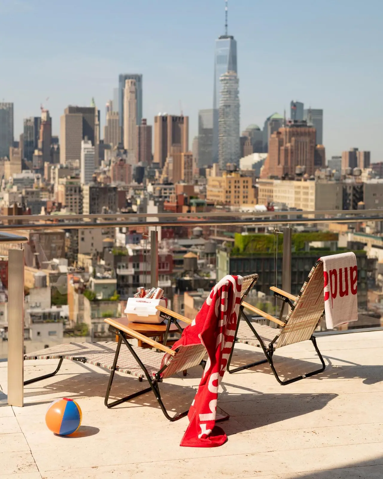 A rooftop terrace with two lounge chairs, red towels and a beach ball overlooks a Manhattan skyline that includes One World Trade Center.