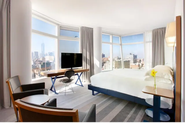 A bedroom with a king bed, a wooden desk with a flat-screen television, armchairs, and large windows showing the city skyline including One World Trade Center.