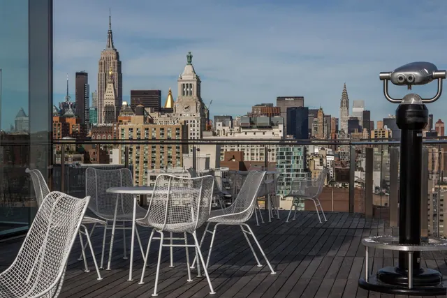 A rooftop terrace with white wire chairs and tables overlooks a city skyline that includes the Empire State Building and the Chrysler Building.