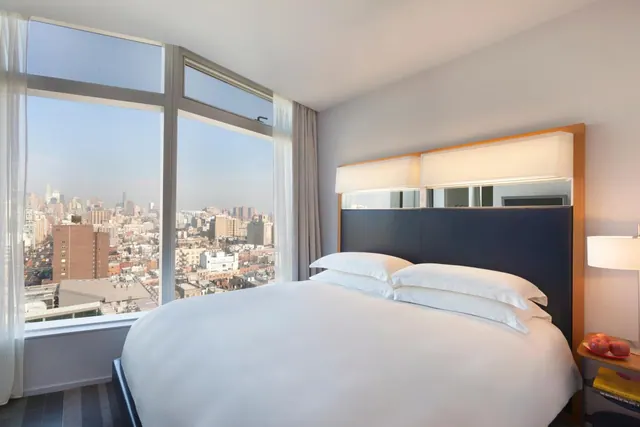 A bedroom with a made king bed, a dark upholstered headboard, and floor-to-ceiling windows overlooking a city skyline.