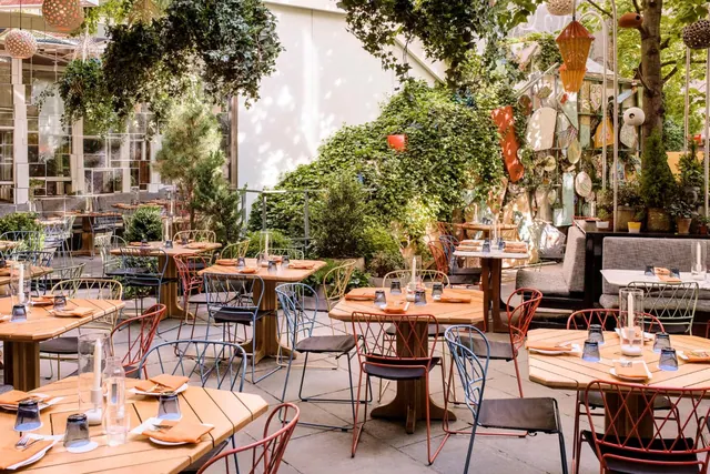 An outdoor dining terrace with wooden tables, metal chairs, orange napkins and hanging greenery beneath a leafy canopy.