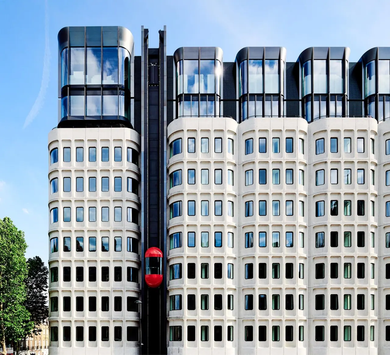 The curved white concrete facade of a multi-storey building with rounded glazed upper floors and a red external lift capsule, against a blue sky.
