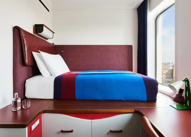 A bedroom with a platform bed under a blue and red striped cover, a red upholstered headboard, a window looking out onto buildings, and a desk with a green lamp.