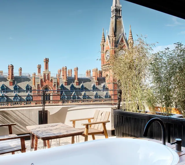 A freestanding bathtub on a rooftop terrace overlooks wooden furniture, bamboo planters and the clock tower of a red-brick Gothic building.