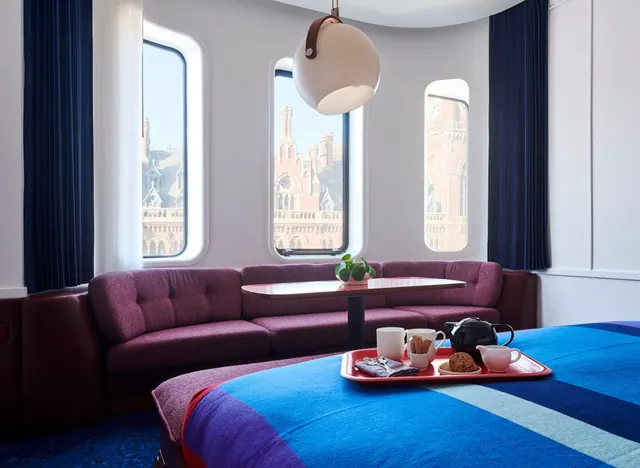 A bedroom with a blue bed cover, a purple sofa and a tea tray on the bed, with windows showing the red-brick St Pancras building outside.