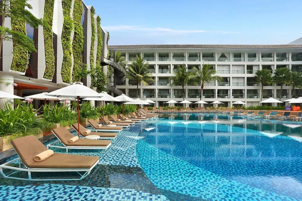 A large swimming pool with blue mosaic tiles, rows of loungers and white parasol umbrellas, with a multi-storey hotel building and palm trees behind.