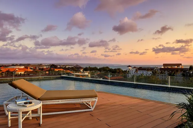 A rooftop pool with a cushioned sun lounger and a side table holding a book and sunglasses, with rooftops and the sea visible under a sunset sky.