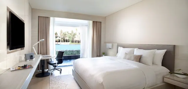 A bedroom with a king bed, a wall-mounted television, a desk and chair, and glass doors looking out onto a pool and palm trees.