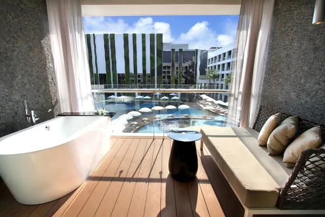 A freestanding bathtub and a cushioned daybed sit on a timber deck, with a glass railing overlooking a pool with umbrellas and loungers.