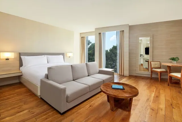 A hotel bedroom with a made king bed, a grey sofa, a round wooden coffee table, and a tall window looking out onto trees.