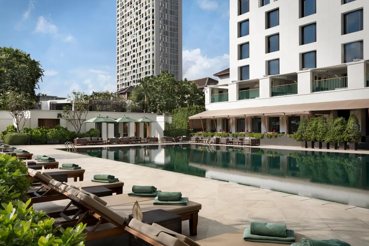 A long swimming pool lined with teak sun loungers topped with green towels, green parasol umbrellas and a white hotel building beside it.
