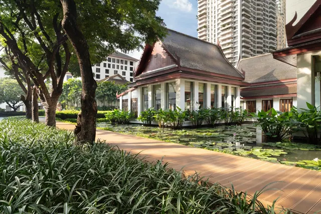 A wooden walkway runs past a lily pond and a traditional Thai-style pavilion, with a tall residential tower behind it.