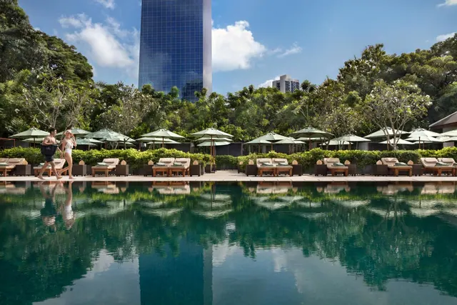 A large pool reflects trees and a tall glass tower, with rows of sun loungers under green umbrellas along the edge and two people walking beside the water.