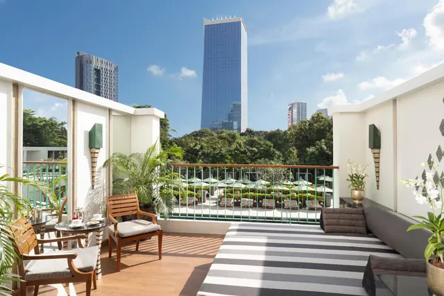 A terrace with wooden chairs, a small table set for tea, a striped daybed and potted plants overlooks a pool with umbrellas, trees and a tall glass tower.