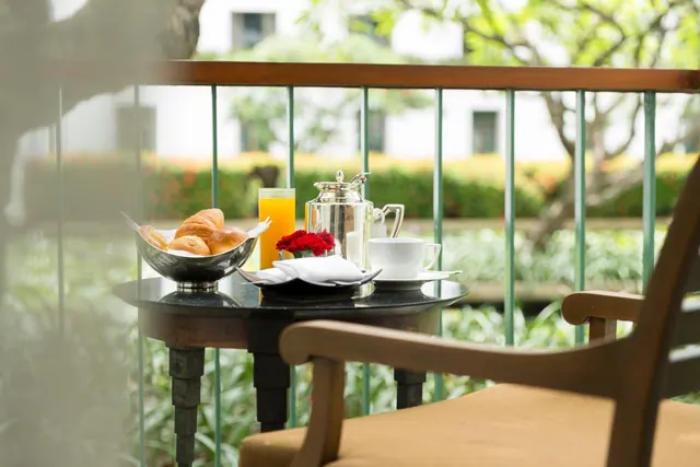 A breakfast setting with a bread basket, orange juice, a silver teapot and a white cup sits on a small round table on a balcony overlooking a garden.