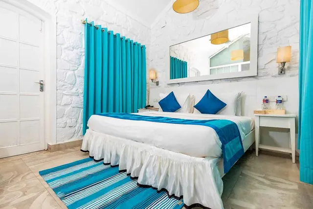 A bright bedroom has a double bed with blue accents, white textured walls, turquoise curtains, and a large wall mirror.