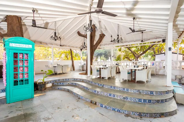 An open-sided dining area has white tables and chairs, patterned tiled steps, hanging fans, and trees beyond the railing.