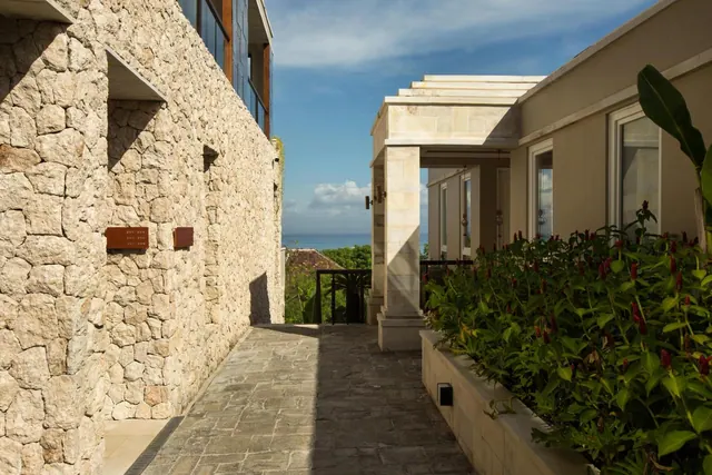 A stone-paved walkway runs between a stone wall and a cream building, with red-flowered plants in a planter and the sea visible in the distance.