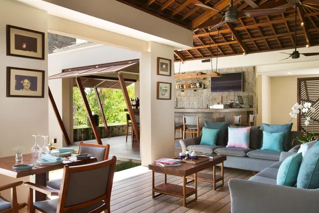 An open-plan living area with grey sofas, a wooden coffee table, a dining table and a stone-walled bar with a wall-mounted television, with greenery visible through the open side.