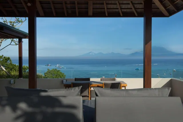 A covered terrace with grey loungers and a wooden dining table overlooks the sea, with boats and distant mountains visible.
