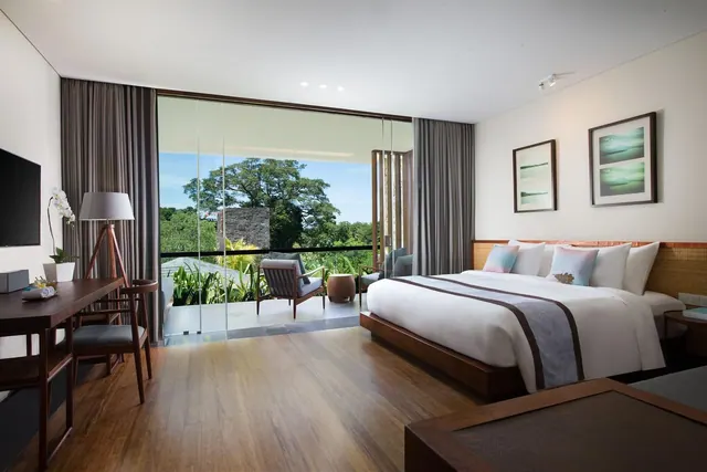 A bedroom with a king bed, a wooden work desk and a wall-mounted television, with floor-to-ceiling glass doors opening onto a balcony with chairs and tall trees beyond.