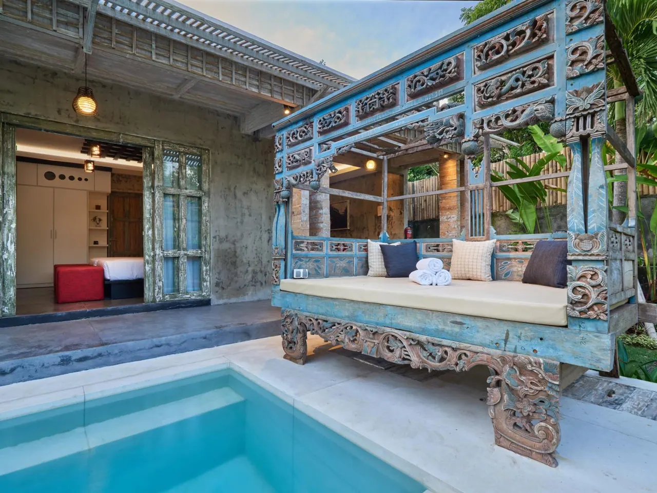 A carved blue wooden daybed with cushions and folded towels sits beside a small pool, with an open doorway into a bedroom behind.