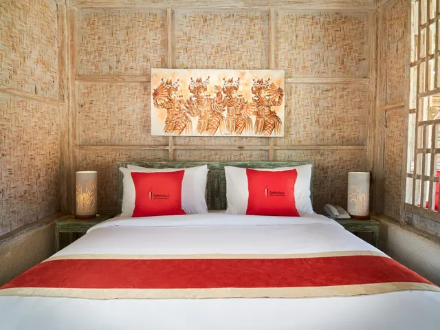 A double bed with red and white bedding and red cushions sits below a painting of dancers on a woven bamboo wall, with a lamp and a telephone on each bedside table.