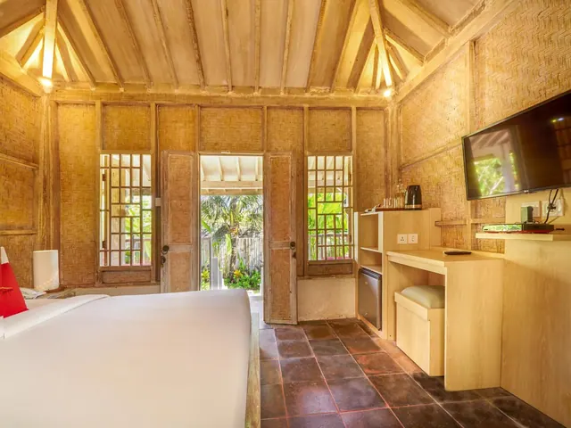 A bedroom with a large white bed, woven bamboo walls, a wooden beamed ceiling, a desk, a wall-mounted television, and an open door onto a garden with palm trees.
