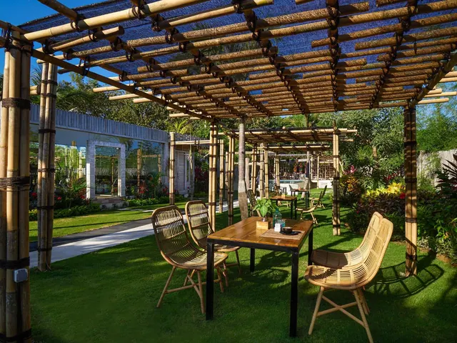 A bamboo pergola shelters a wooden dining table and rattan chairs on a grass lawn beside a pathway and landscaped plants.