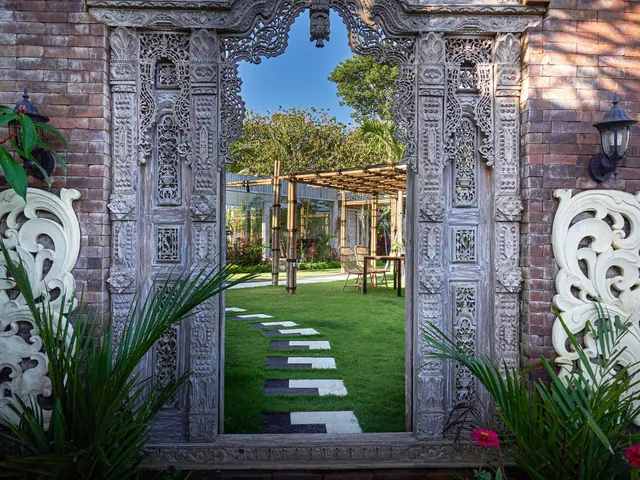A carved wooden gateway in a brick wall frames a lawn with stepping stones, a bamboo pergola, and a table with chairs beyond.