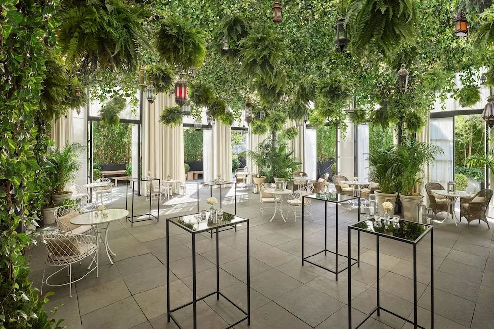 A covered garden terrace with hanging ferns, lanterns, white metal chairs and tables, and glass-top pedestal tables on a stone floor, with glass doors opening onto greenery.
