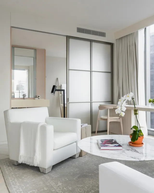 A white armchair, a round marble coffee table with books and an orchid, and a wood desk with chair sit in a bright hotel room, beside frosted sliding doors and a partly visible bathroom.