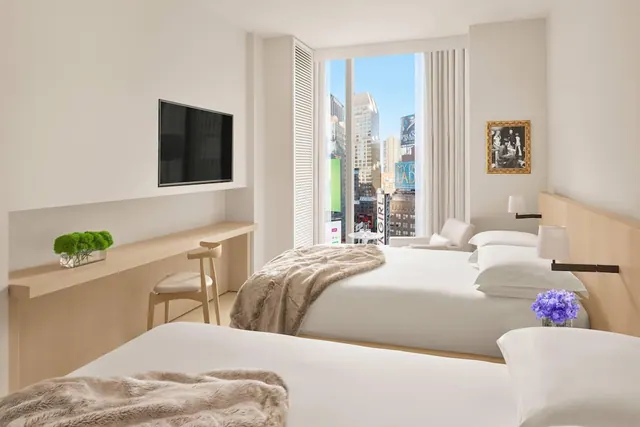 A hotel bedroom with two beds, a wall-mounted television, a wooden desk with a chair, and a tall window overlooking city buildings and billboards.