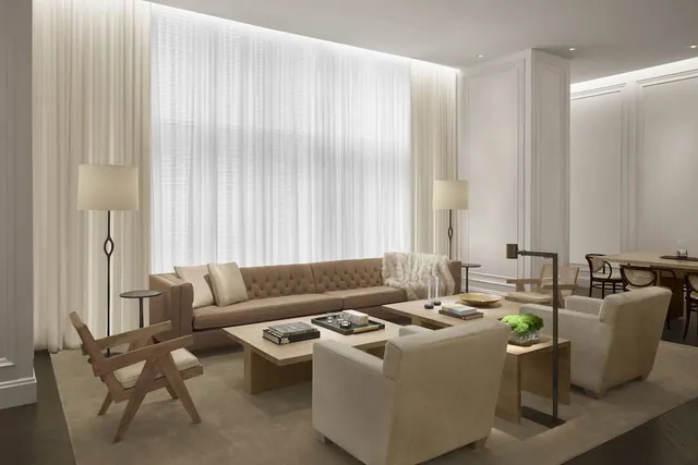 A staged living area with a tufted sofa, upholstered armchairs, wooden coffee tables, floor lamps and a large window covered with sheer curtains.