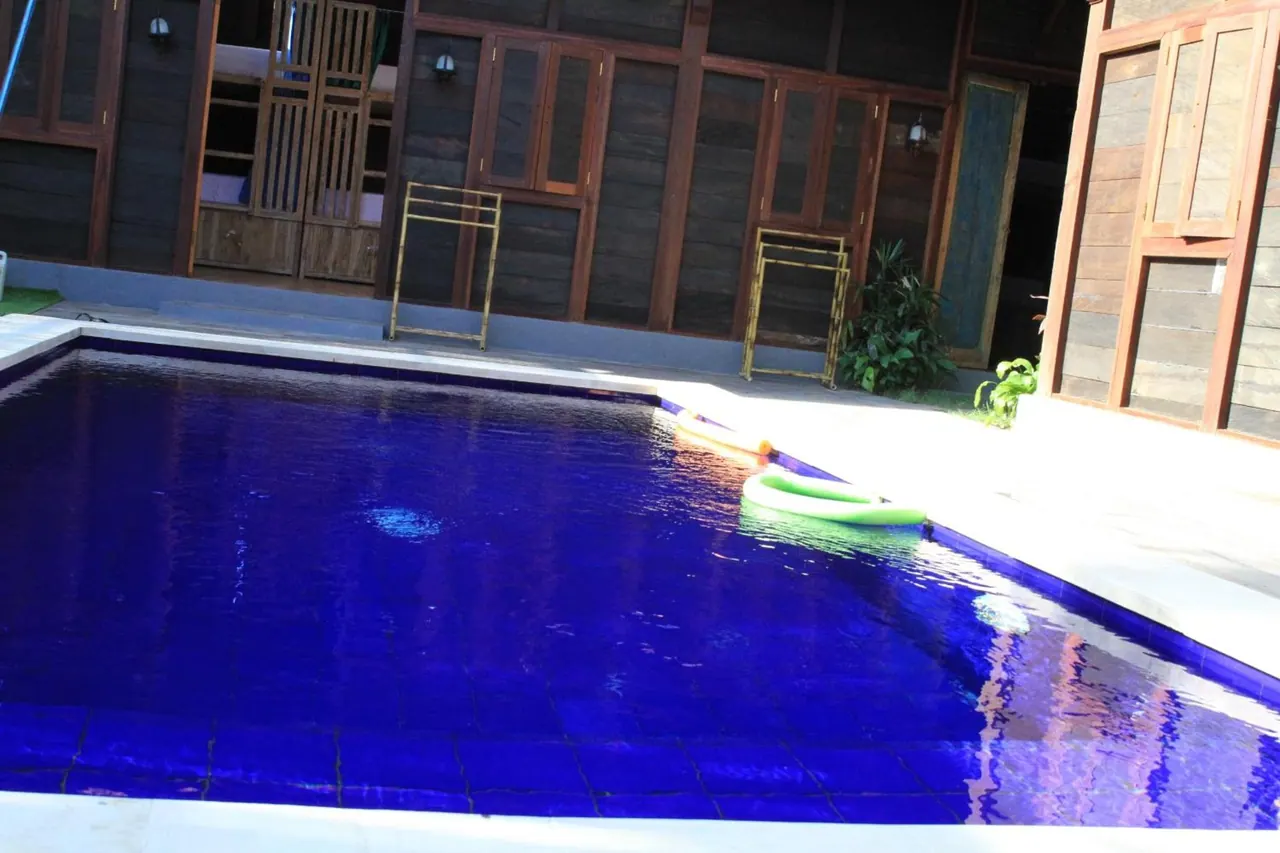 A rectangular blue-tiled swimming pool with a green pool noodle floating beside a wooden building with towel racks.