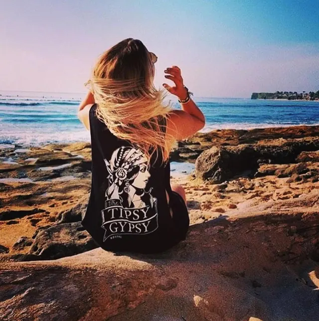 A woman with long hair, seen from behind in a black tank top with printed text, sits on a rocky shore looking out at the sea.