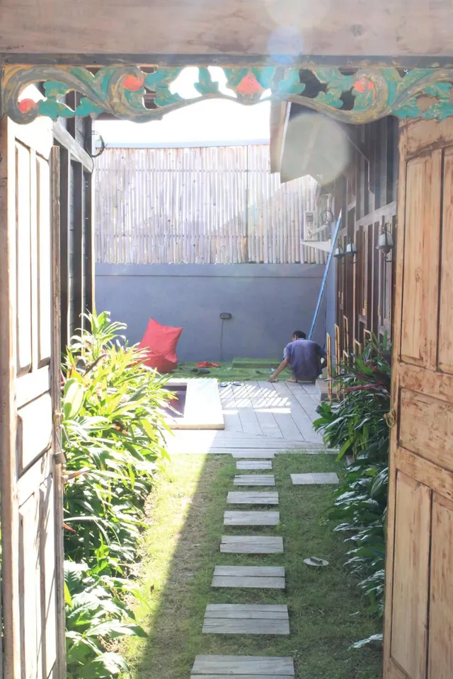A carved wooden gateway frames a grassy garden path with stepping stones, with a worker kneeling near a red cushion beside a grey wall and bamboo fence.