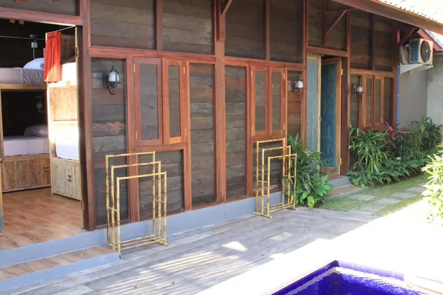 Exterior of a timber-clad building with a wooden deck, an open doorway showing bunk beds inside, and a small blue pool at the lower right.