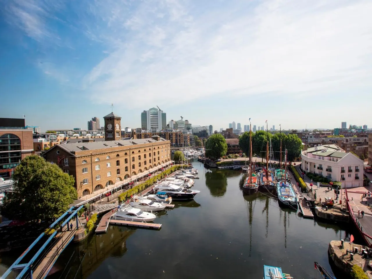 Elevated view over a dock with moored motor yachts and sailing boats toward a brick warehouse with a clock tower and distant city towers under a blue sky.