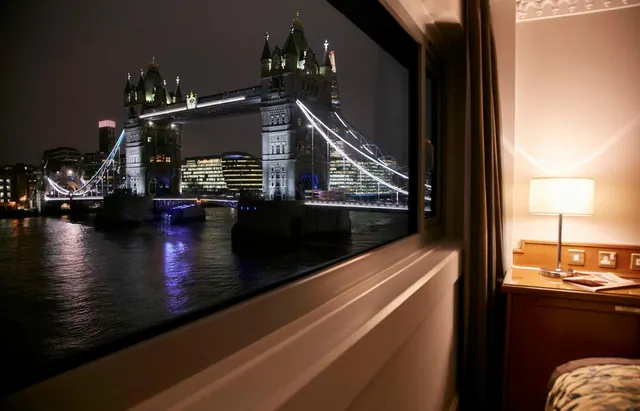 A night view through a hotel room window of Tower Bridge over the River Thames, with a bedside lamp and nightstand in the room.